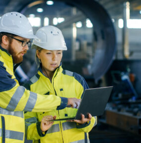 Male and Female Industrial Engineers in Hard Hats Discuss New Project while Using Laptop. They Make Showing Gestures.They Work in a Heavy Industry Manufacturing Factory.