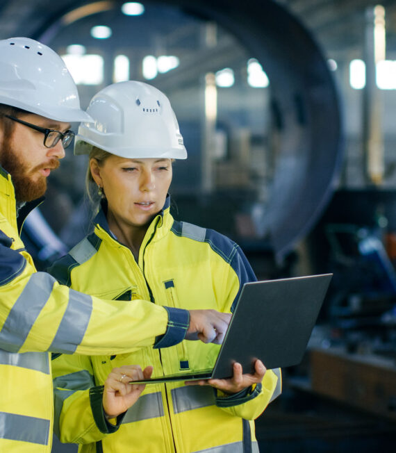 Male and Female Industrial Engineers in Hard Hats Discuss New Project while Using Laptop. They Make Showing Gestures.They Work in a Heavy Industry Manufacturing Factory.